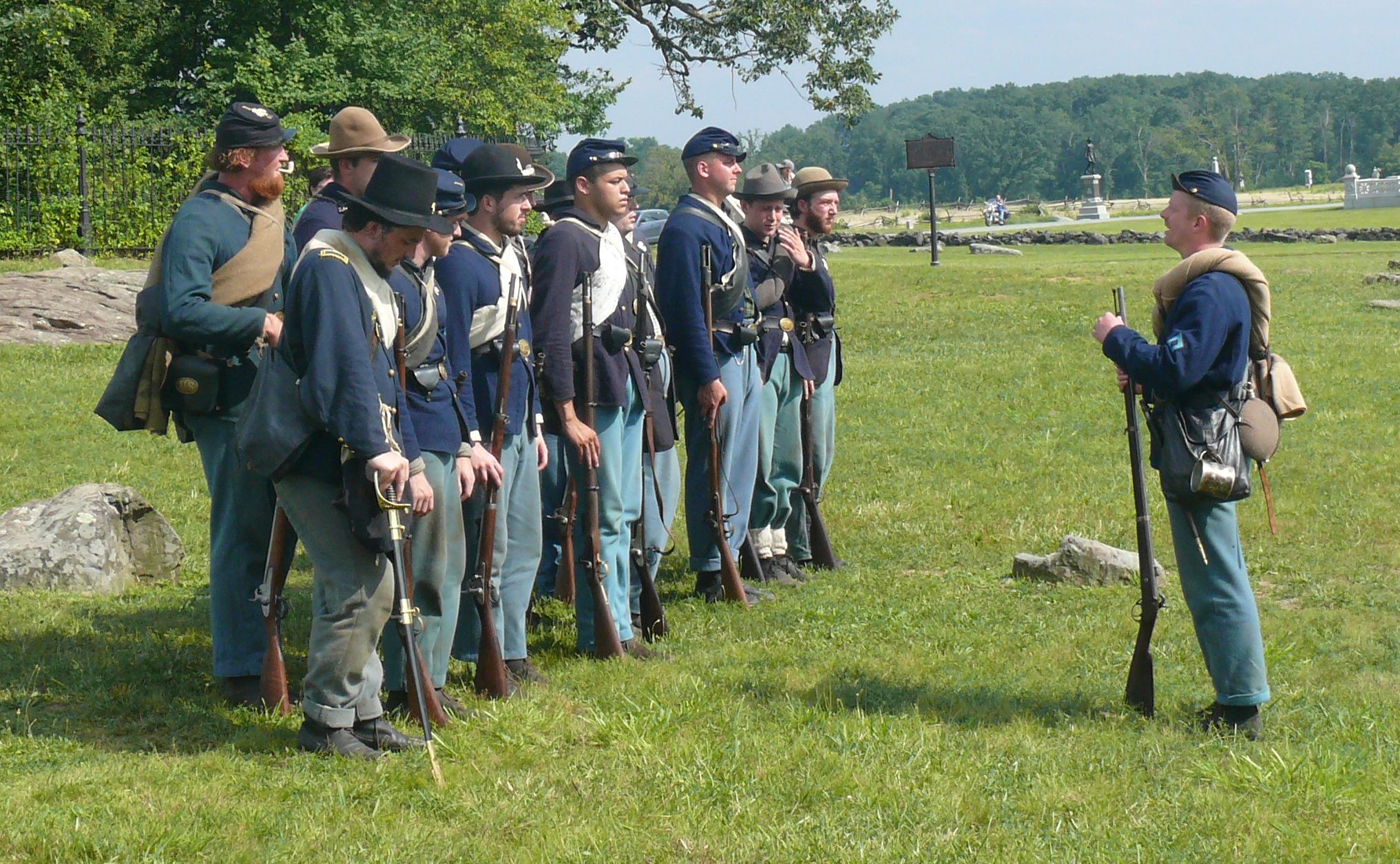 Eine Gruppe Männer in unterschiedlichen blauen Nordstaaten-Uniformen steht auf einer Wiese vor Bäumen; ihnen gegenüber steht ein ebenso gekleideter Offizier und hält eine Ansprache