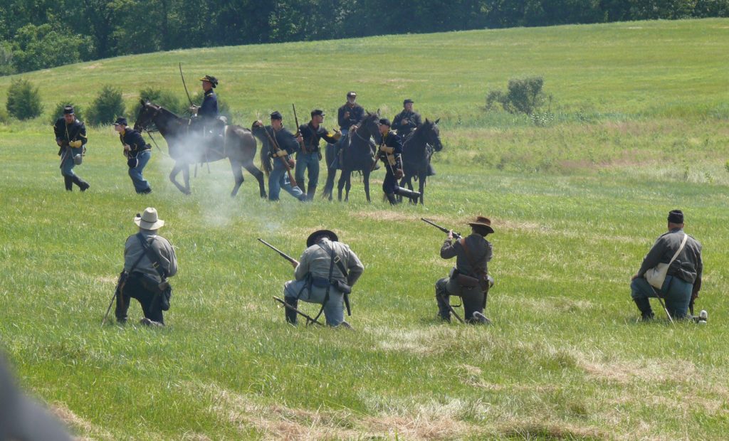 Fußsoldaten in grauer Konföderierten-Uniform knien im Vordergrund vor einer Gruppe blau uniformierter Unionssoldaten